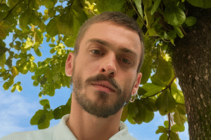 Retrato do ator Felipe Selau sob a sombra de uma árvore, com fundo de folhas verdes e céu azul, imagem otimizada por IA.