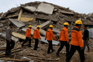 Equipes de busca caminham entre escombros de uma construção colapsada na Indonésia. Imagem gerada por IA.