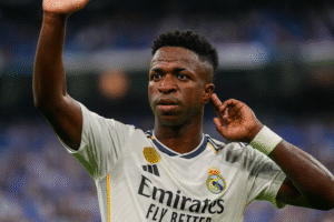 Vinícius Júnior em campo com o uniforme do Real Madrid, gesto atento com a mão próxima ao ouvido, durante partida simbólica. Imagem ilustrativa gerada por inteligência artificial.