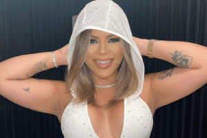 Mulher sorridente com capuz branco e roupa clara com brilhos, posando com os braços levantados; imagem redimensionada por inteligência artificial.