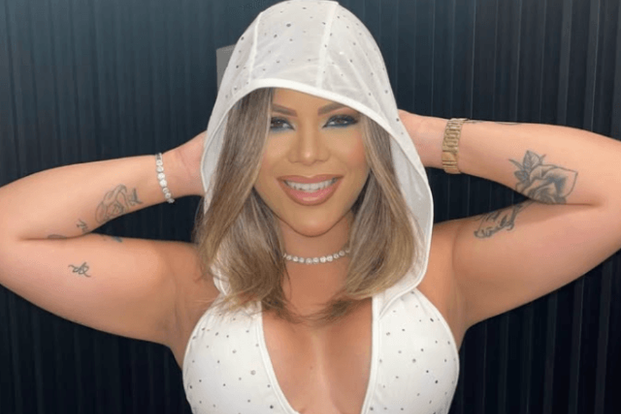 Mulher sorridente com capuz branco e roupa clara com brilhos, posando com os braços levantados; imagem redimensionada por inteligência artificial.