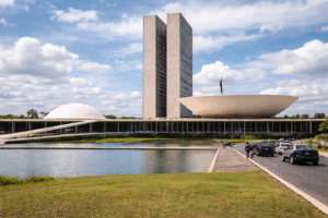 Vista externa do Congresso Nacional, em Brasília, com as torres do Legislativo, cúpulas do Senado e da Câmara, espelho d’água, carros e pedestres em um dia parcialmente ensolarado.
