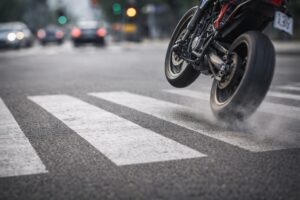 Imagem ilustrativa mostra motocicleta empinada atravessando faixa de pedestres em cruzamento urbano durante o dia.