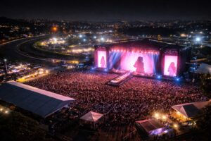 Vista aérea noturna do Autódromo de Interlagos com palco iluminado em rosa e público durante festival; imagem ilustrativa gerada por IA.