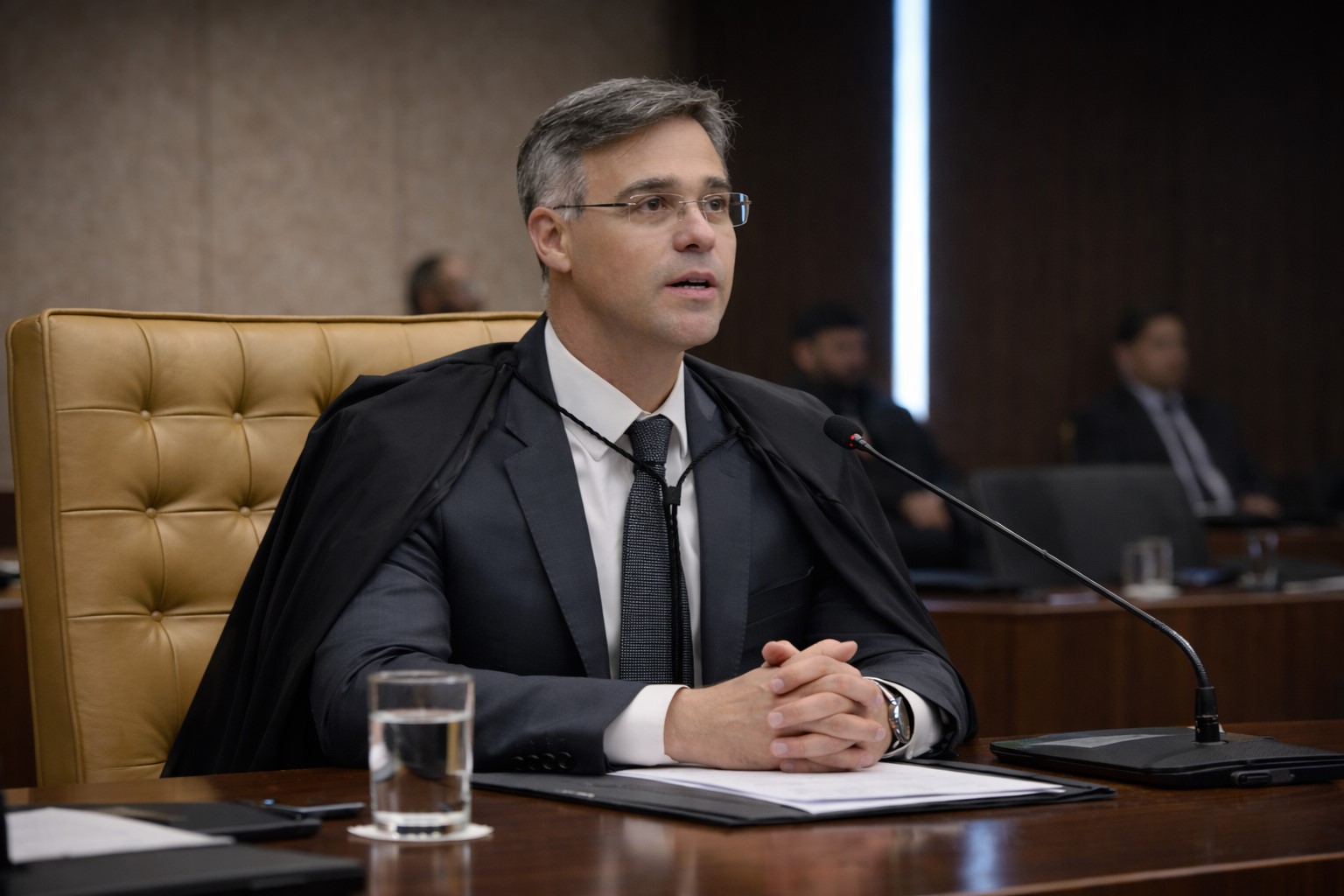 Imagem gerada por IA do ministro André Mendonça sentado em mesa do STF, de meio perfil, aguardando discurso em ambiente institucional.