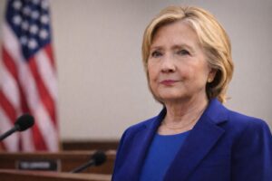 Retrato ilustrativo de Hillary Clinton em ambiente institucional com bandeira dos Estados Unidos ao fundo, imagem gerada por IA para contextualização jornalística.