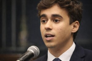 Imagem gerada por IA de Nikolas Ferreira discursando em ambiente que remete à Câmara dos Deputados, com microfone e fundo institucional desfocado.