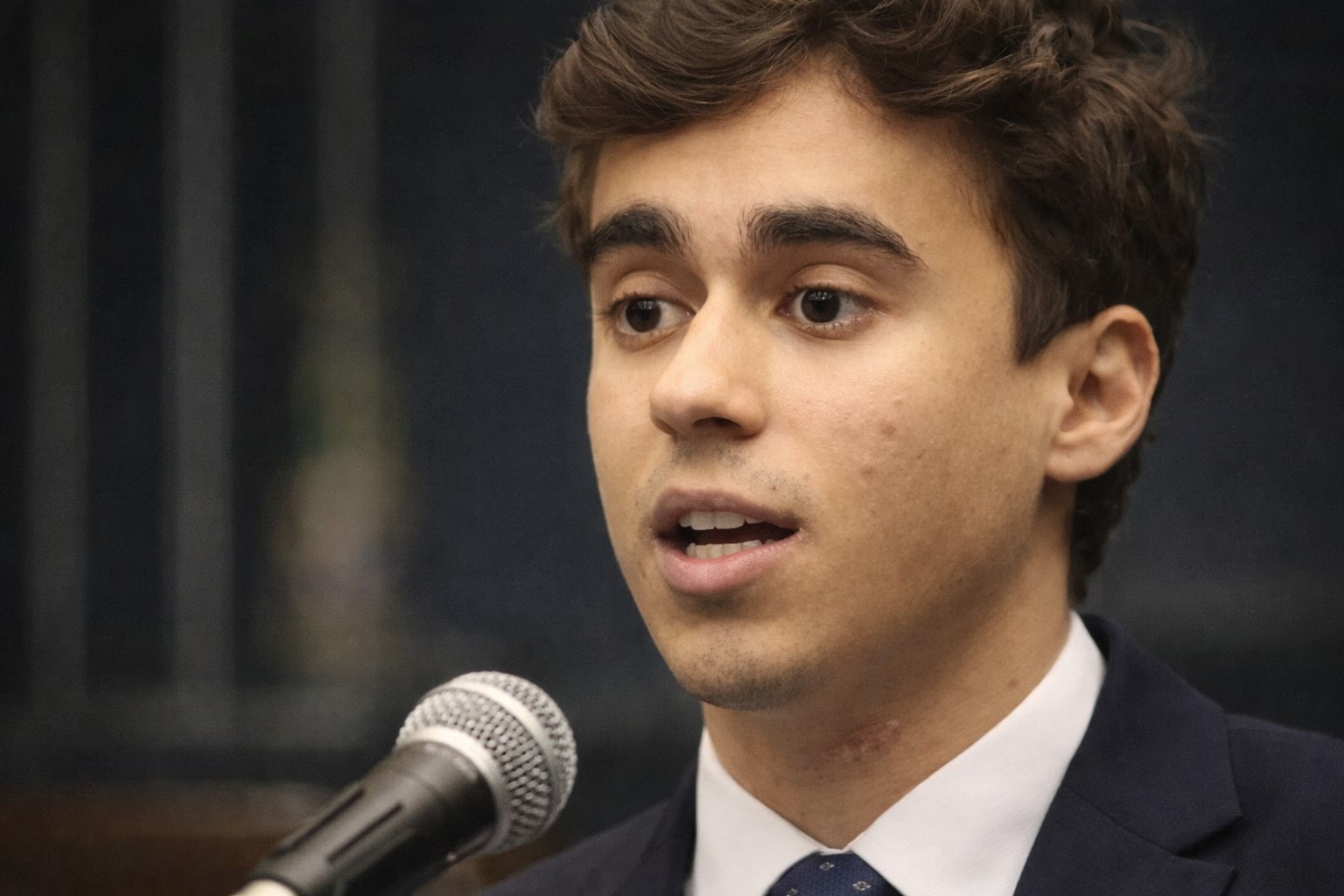 Imagem gerada por IA de Nikolas Ferreira discursando em ambiente que remete à Câmara dos Deputados, com microfone e fundo institucional desfocado.