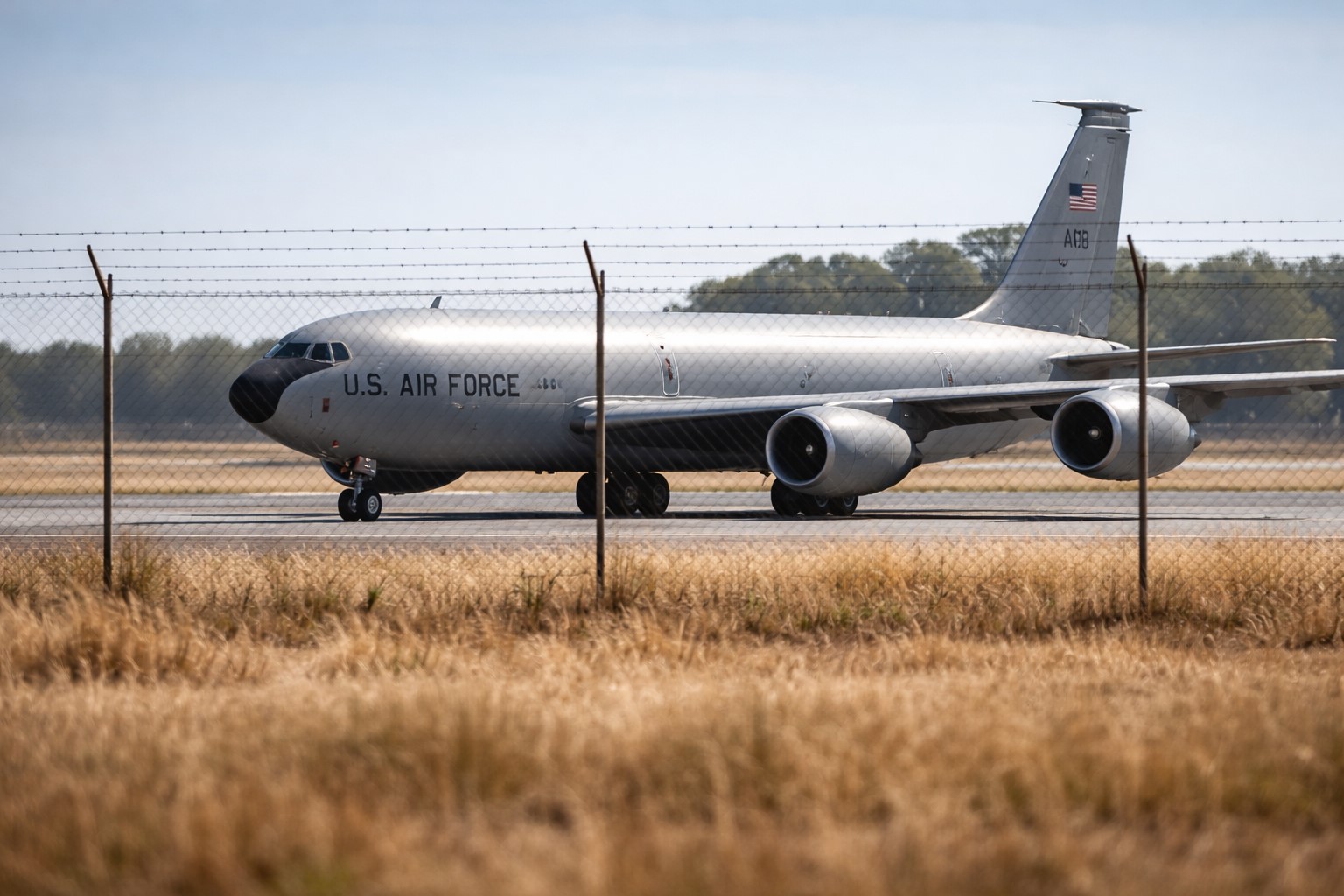 Ilustração fotográfica ultra-realista mostra um avião militar de reabastecimento modelo KC-135 estacionado em pista de aeroporto, atrás de alambrado, em cenário diurno.