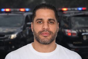 Retrato de Luiz Phillipi Machado de Moraes Mourão, conhecido como “Sicário”, em primeiro plano, com viaturas policiais desfocadas ao fundo representando escolta policial.