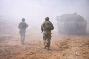 Soldados israelenses caminham em terreno arenoso durante operação militar com veículo blindado ao fundo.