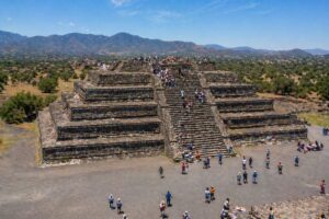 Ilustração fotográfica ultra-realista mostra uma das pirâmides de Teotihuacán, no México, em cena diurna com turistas distribuídos pela estrutura.