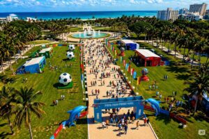 Imagem aérea de festival de futebol com público e telões em parque litorâneo